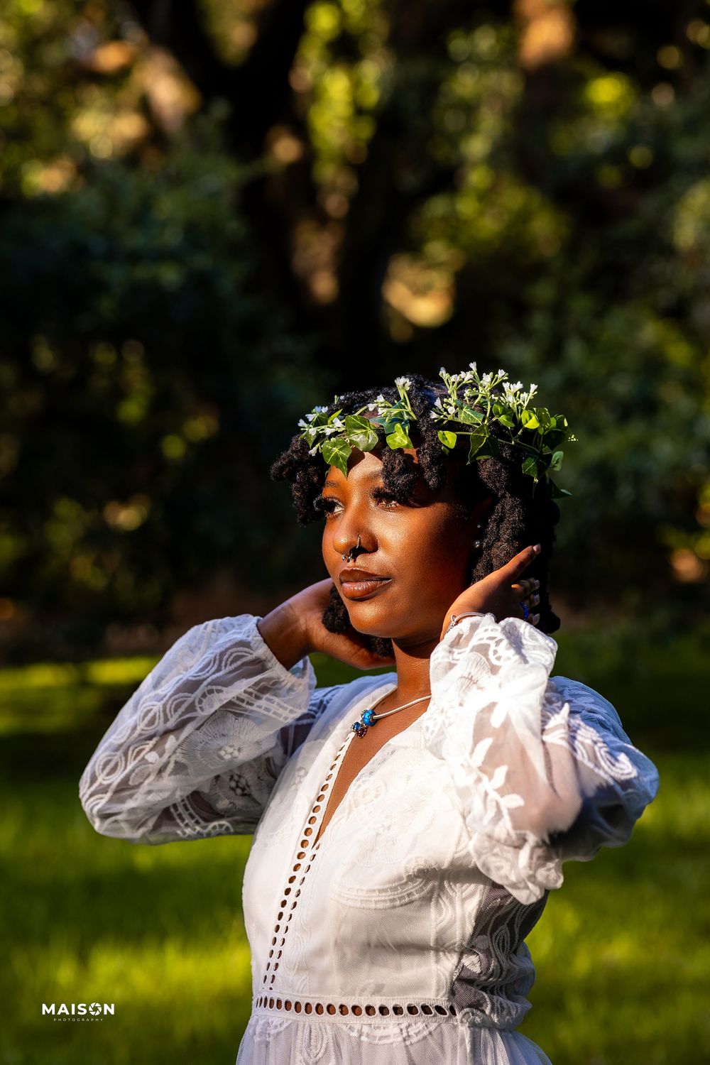A person wearing a white lace dress and floral crown poses in a sunlit garden setting at golden hour.