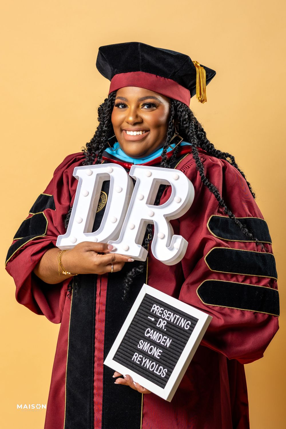 Graduate in burgundy academic regalia holds large 'DR' letters and sign while standing against peach background.