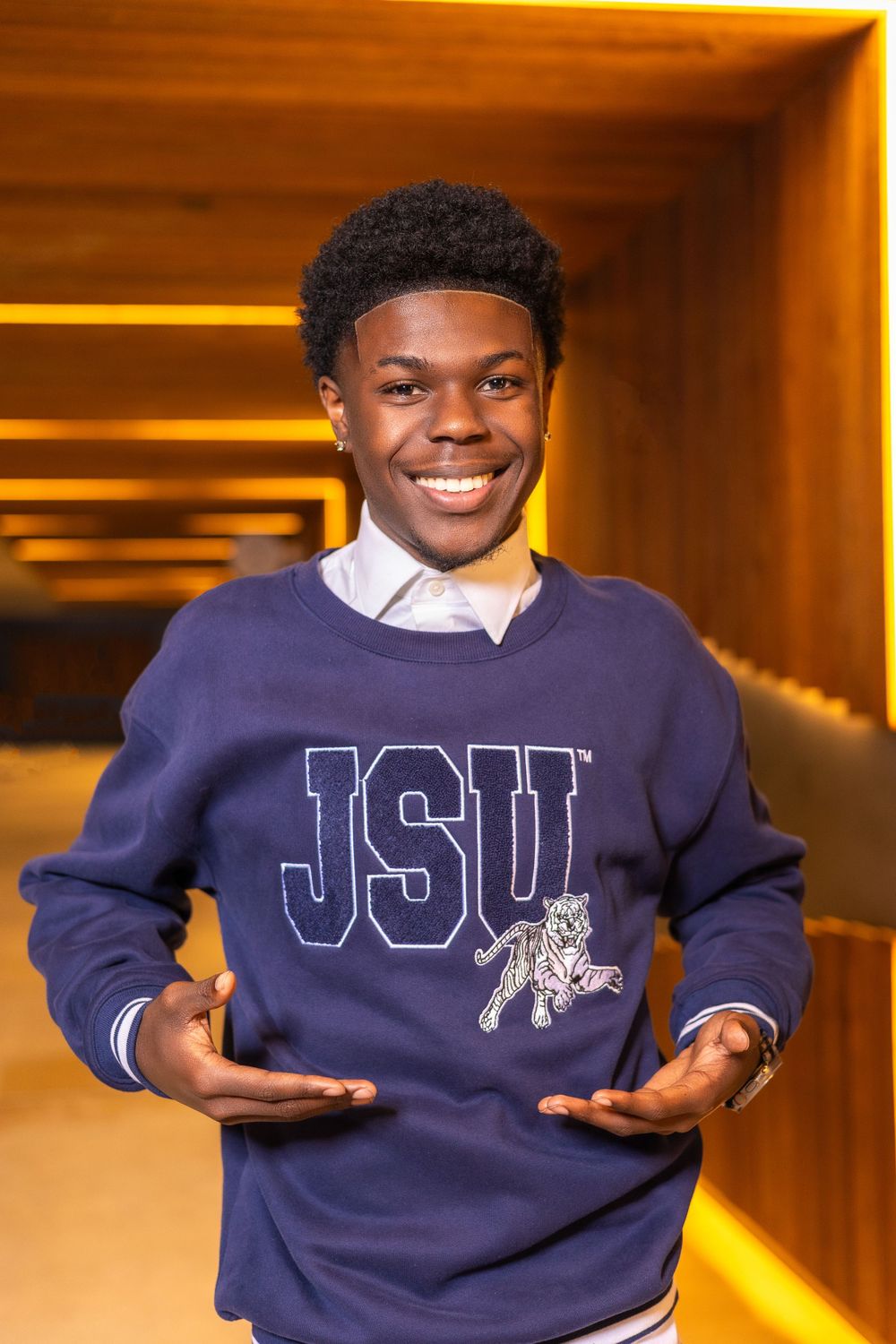 Smiling student wearing a navy JSU collegiate sweater and white collared shirt stands in a warmly lit hallway.