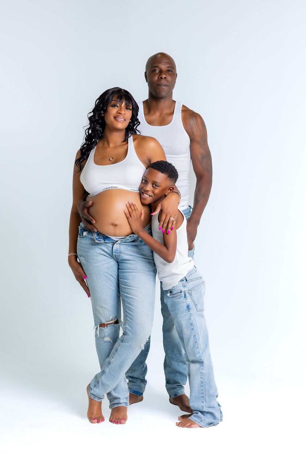 A family poses together against a white backdrop, with a pregnant woman in ripped jeans and two others in casual attire.