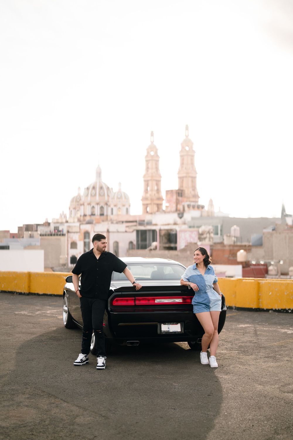 Couple posing with classic black car on rooftop with cathedral towers in background.