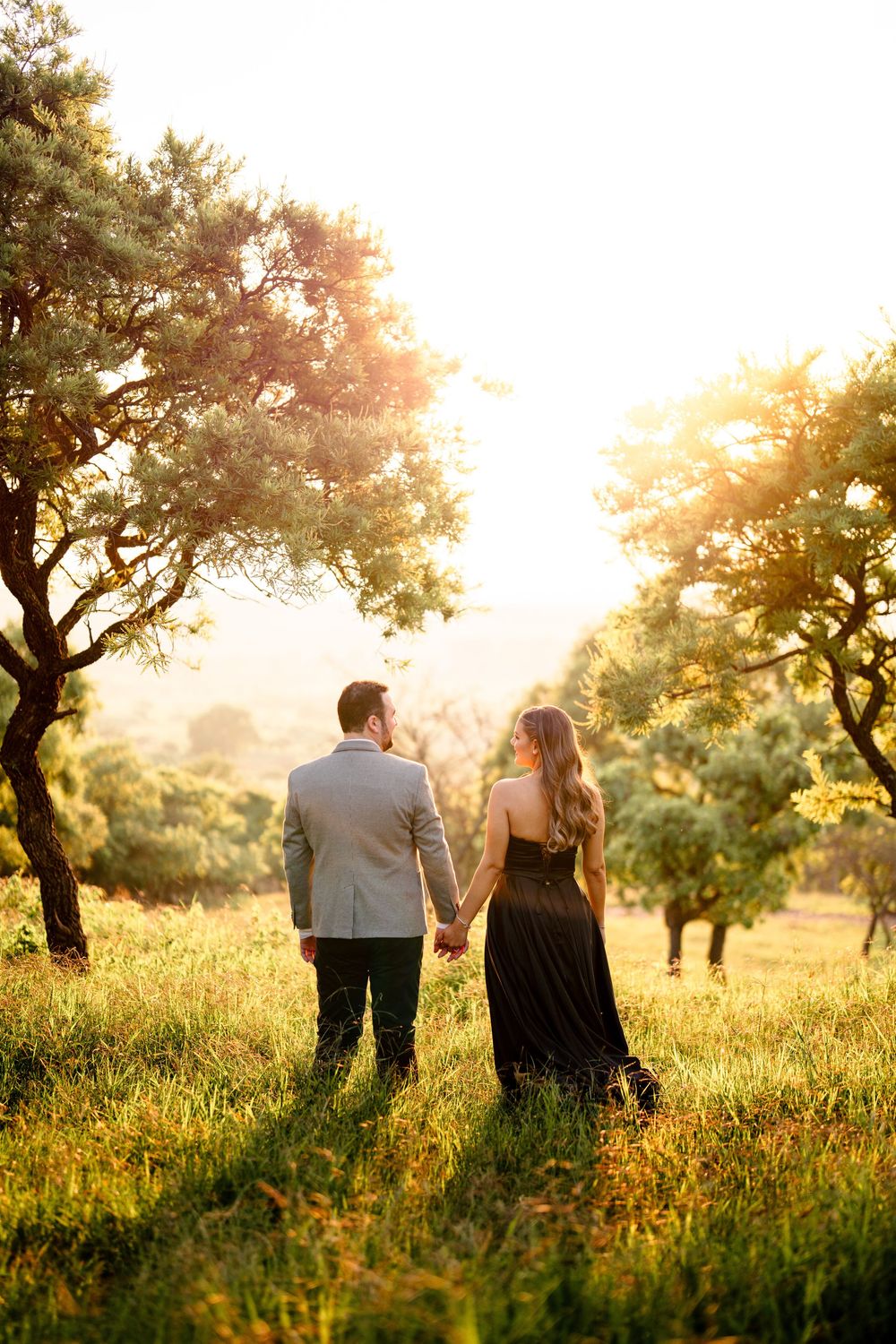 Couple walking through a golden meadow at sunset with trees silhouetted in the background.