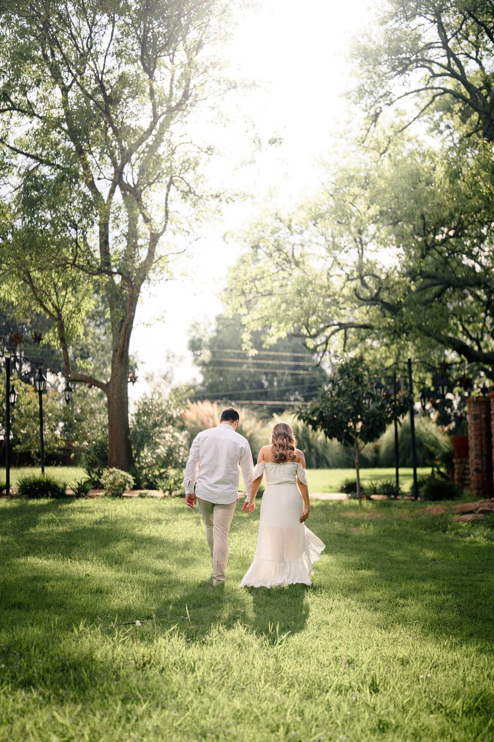 Couple walking hand in hand through a sunlit garden path with lush green trees.