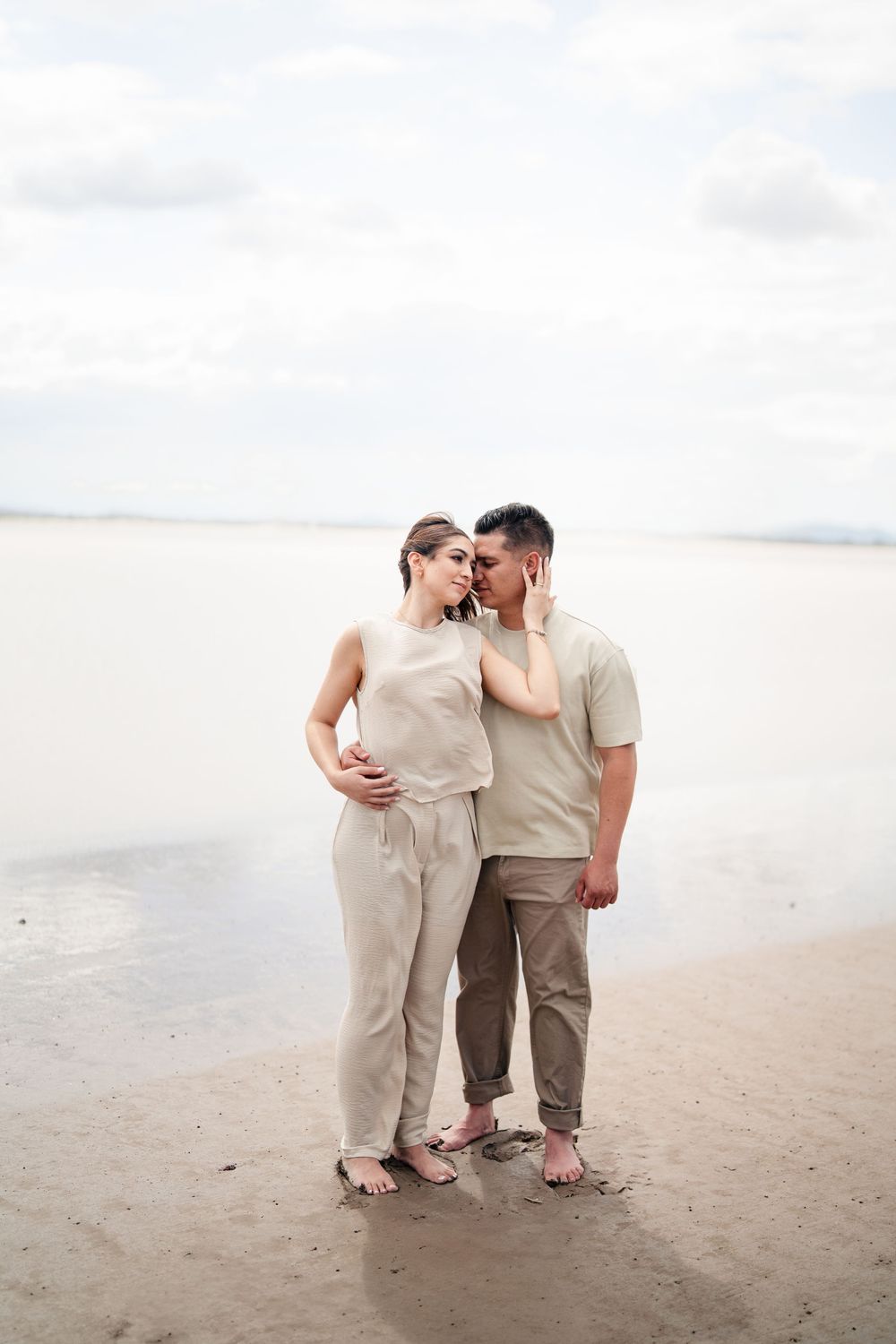 Couple in neutral colored outfits sharing intimate moment on beach.