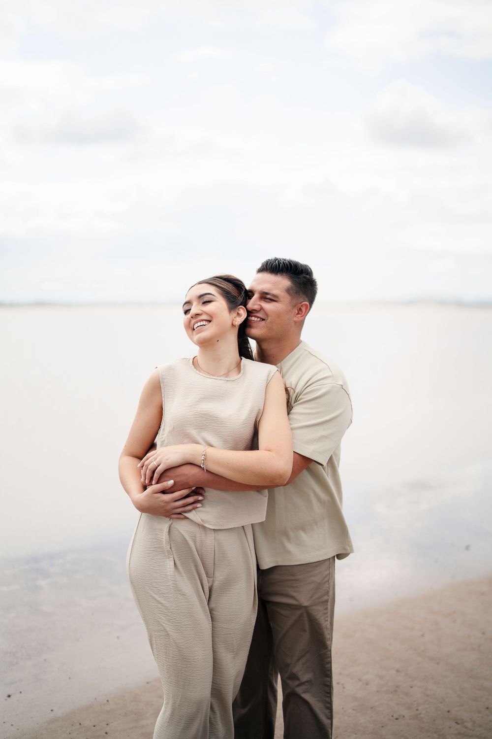 Couple in beige clothing embracing on beach during cloudy day.