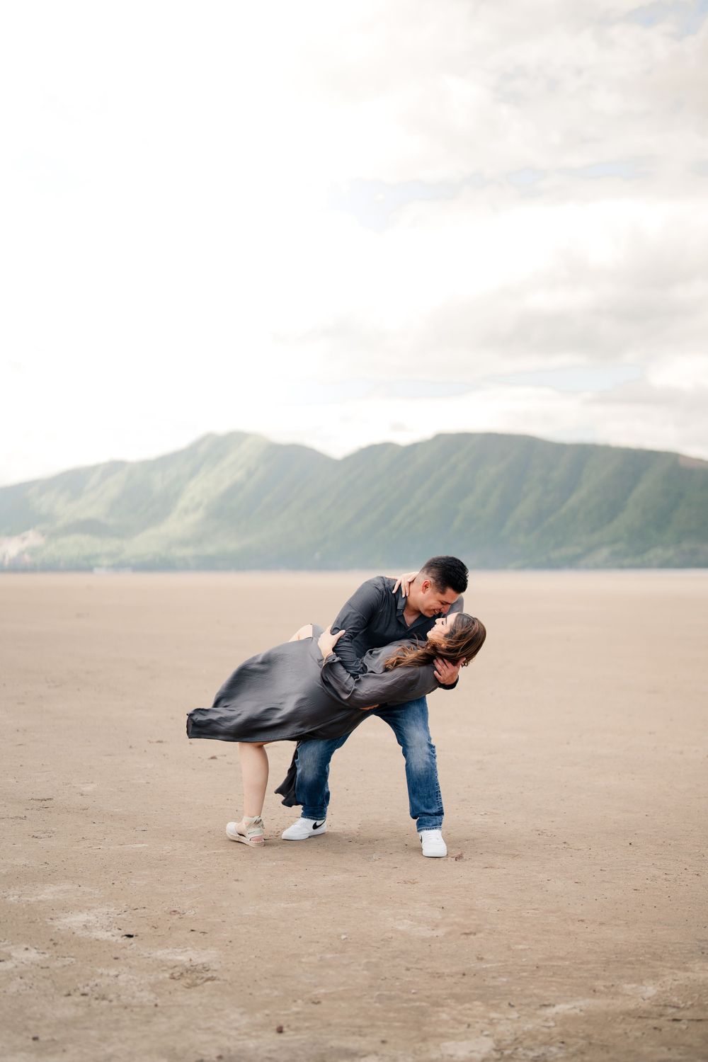 Romantic dip pose on sandy beach with mountains and cloudy sky in background.