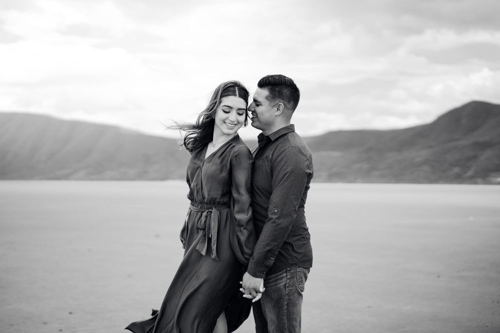 Black and white photo of couple embracing on beach with mountains in distance.