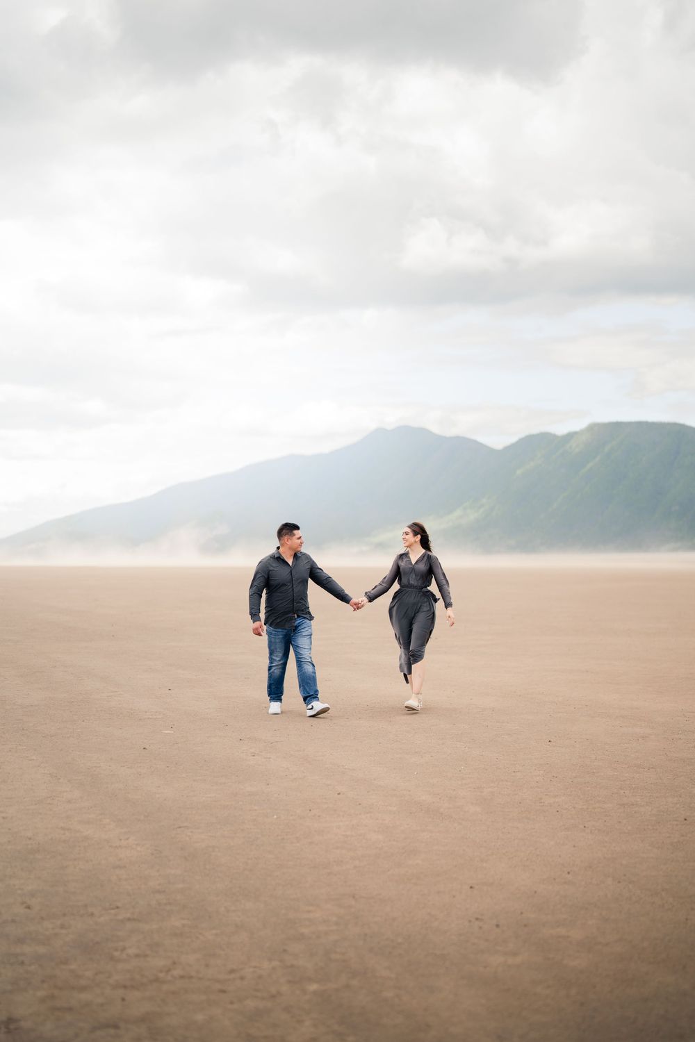 Couple holding hands while running on a vast beach with mountains in the background.