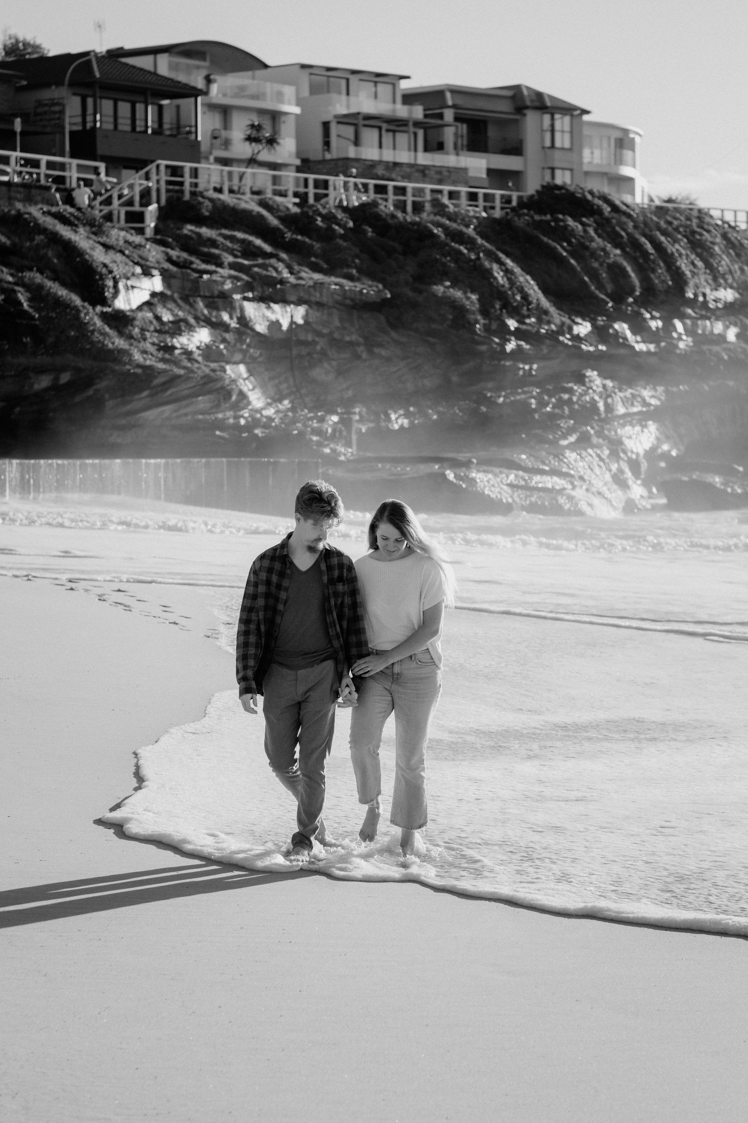 Black and white photo of a couple walking along a beach with coastal homes on cliffs in the background.