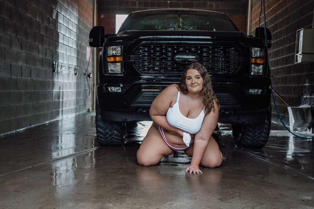 Woman in white swimsuit kneeling while washing a black truck in a garage car wash setting.