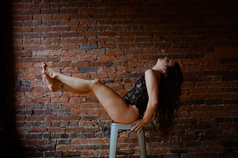 Woman in black lace bodysuit posing on a stool against a rustic exposed brick wall.
