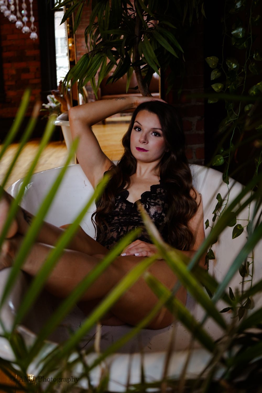 Woman in black lace lingerie relaxing in a bathtub surrounded by tropical indoor plants.