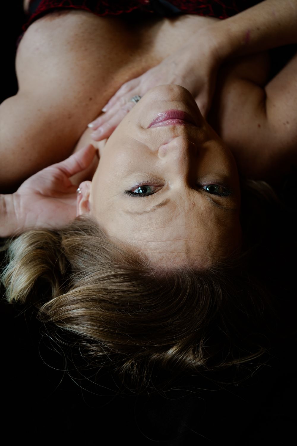 Woman lying back with hands near face, captured in an artistic upside-down portrait with dramatic lighting.