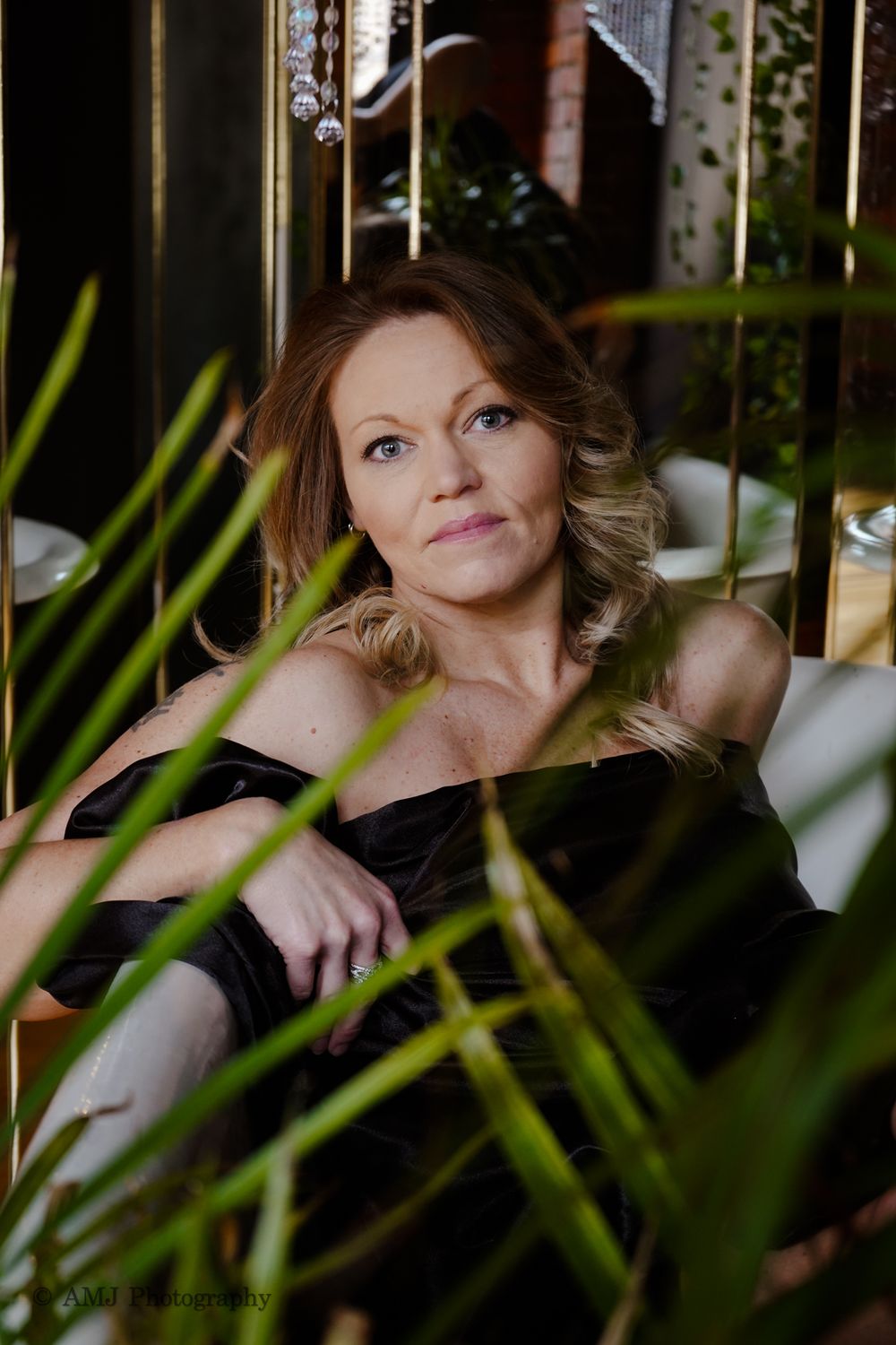 Elegant woman in black off-shoulder dress seated among lush green plants in a stylish interior setting.