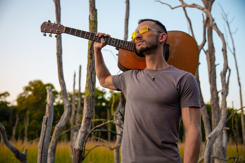 Guitarist in gray t-shirt playing acoustic guitar outdoors among bare trees at sunset.