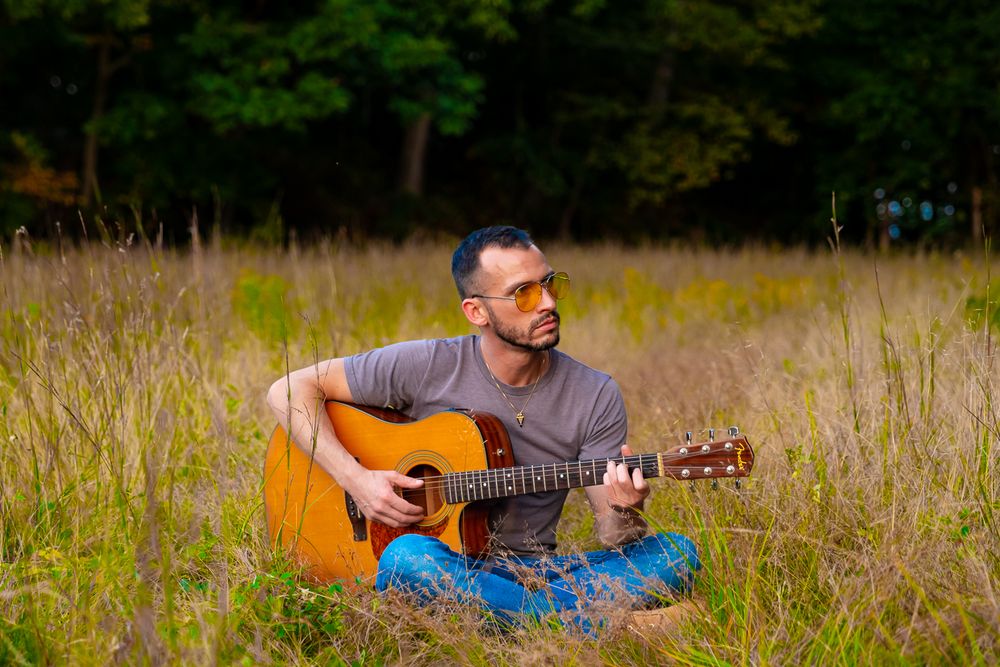 Musician playing acoustic guitar while sitting in tall grass field on sunny day.