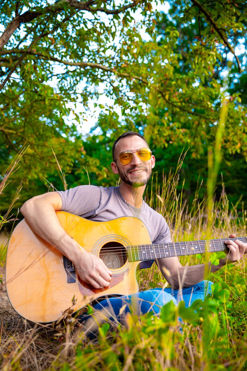 Guitarist playing acoustic guitar outdoors in natural setting with sunlit trees in background.
