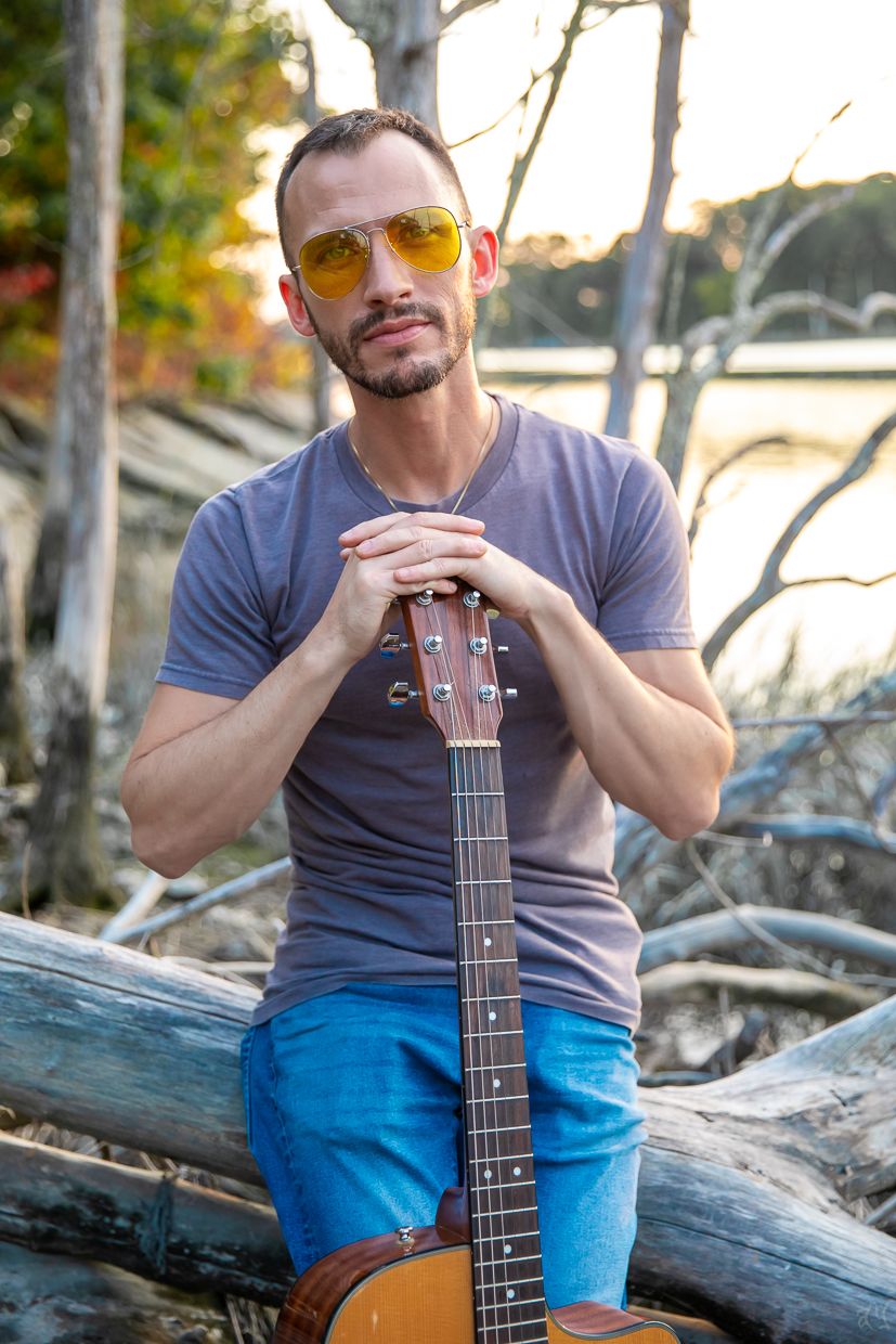 Musician with yellow sunglasses sitting on driftwood holding an acoustic guitar by the water.