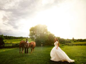 Bride in white gown stands on lush green farm at golden hour with two brown horses grazing nearby.