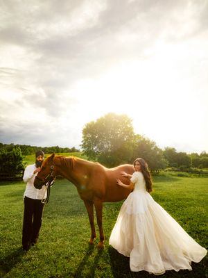 Bride in white gown and groom pose with a chestnut horse in a sunlit green field during a romantic outdoor wedding photo.