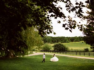 Bride and groom walking hand in hand through lush green gardens with rolling hills and wooden fence backdrop.