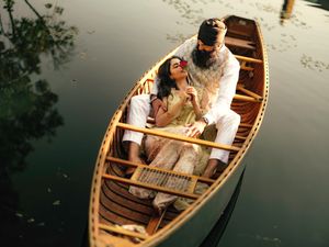 A romantic couple in traditional attire shares an intimate moment in a wooden canoe on a calm reflective lake.