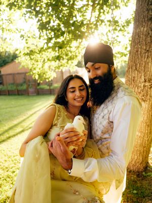 A Sikh couple in traditional attire holding a white dove under a sunlit tree in a garden.
