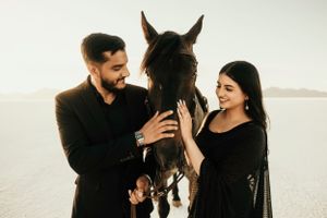 A couple in black outfits poses with their dark horse in a dreamy desert landscape at sunset.