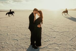 A romantic couple embraces on a salt flat desert with horse riders silhouetted in the background.