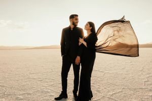 A couple in matching black outfits poses on salt flats with flowing fabric catching the wind.
