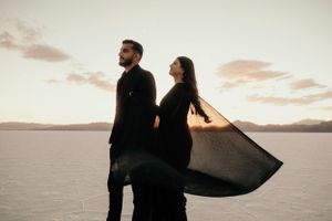 A dramatic portrait of a couple standing against a lake backdrop with flowing black fabric.