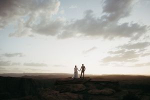A couple in silhouette holds hands on desert cliffs during sunset.