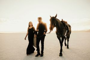 Couple in black attire poses with horse in minimalist desert landscape at sunset.