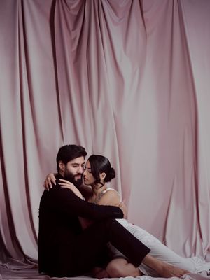 A couple in dark clothing shares an intimate moment while reclining on a pink draped backdrop.