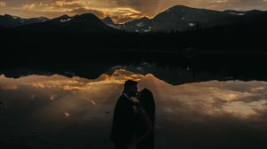 Two silhouettes stand together against a dramatic mountain lake sunset with perfect mirror reflection in the water.