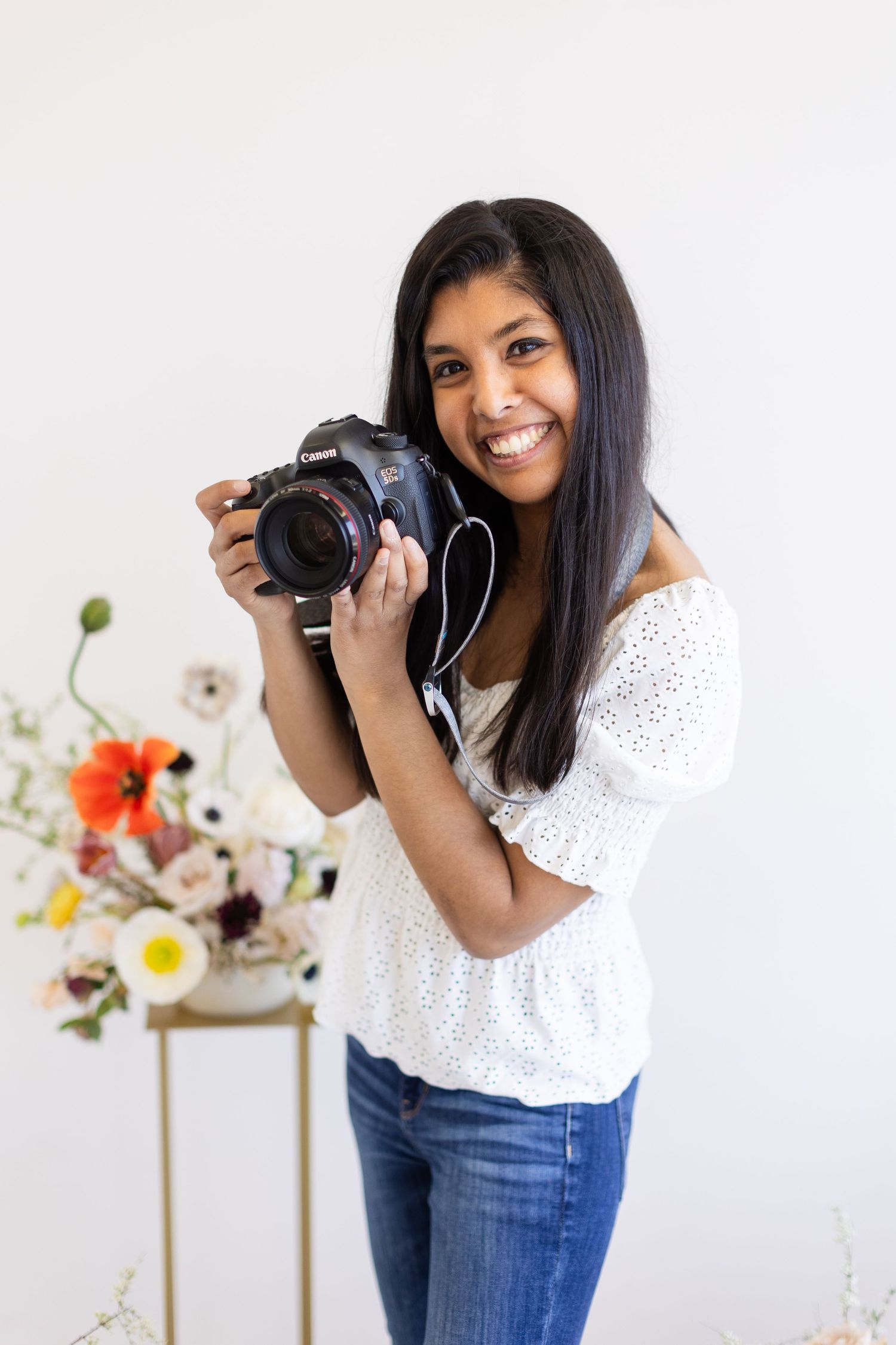 Professional photographer in white eyelet top and jeans holding camera against floral backdrop.