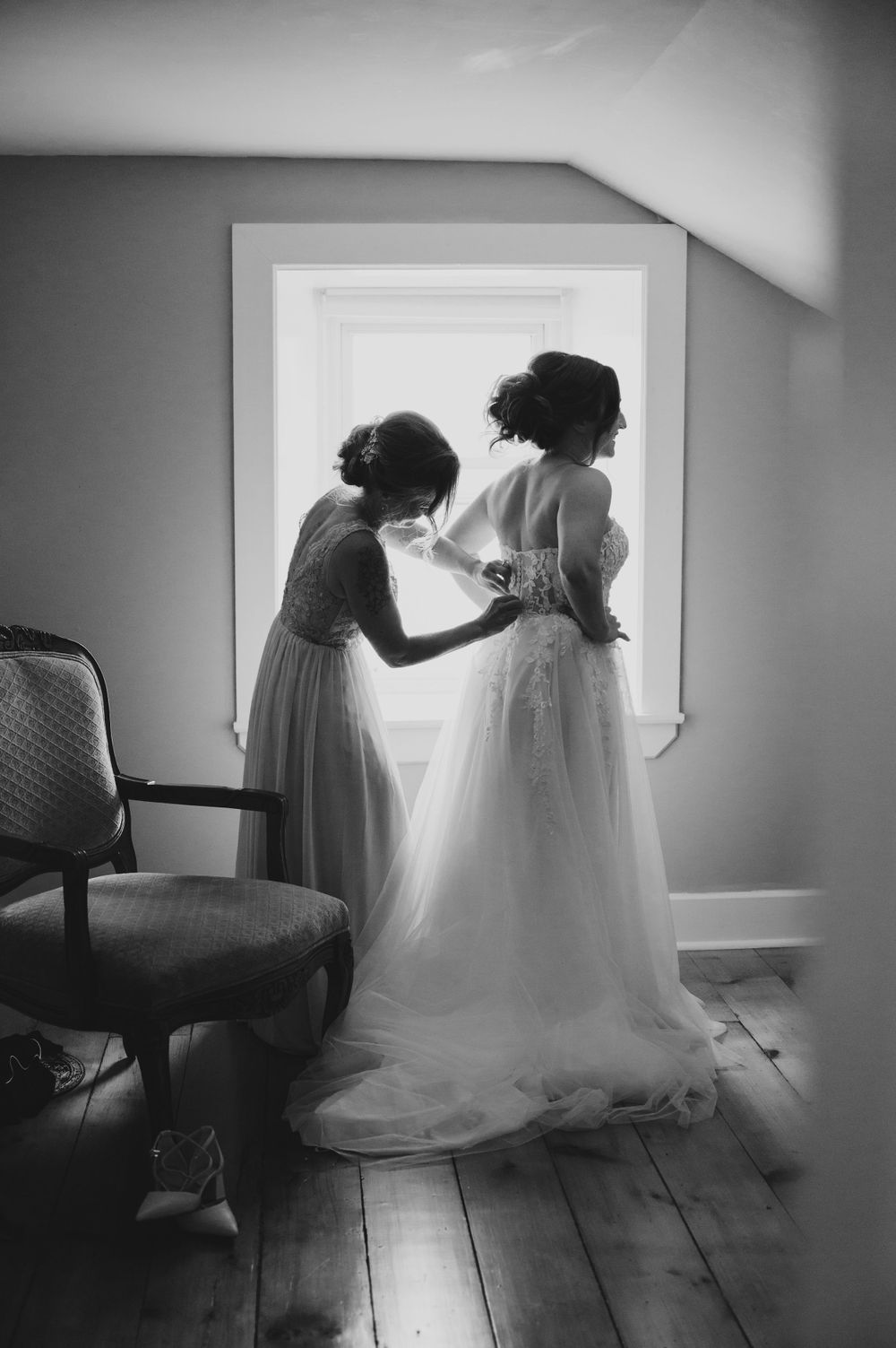 A black and white photo capturing final wedding dress adjustments in a sunlit room.