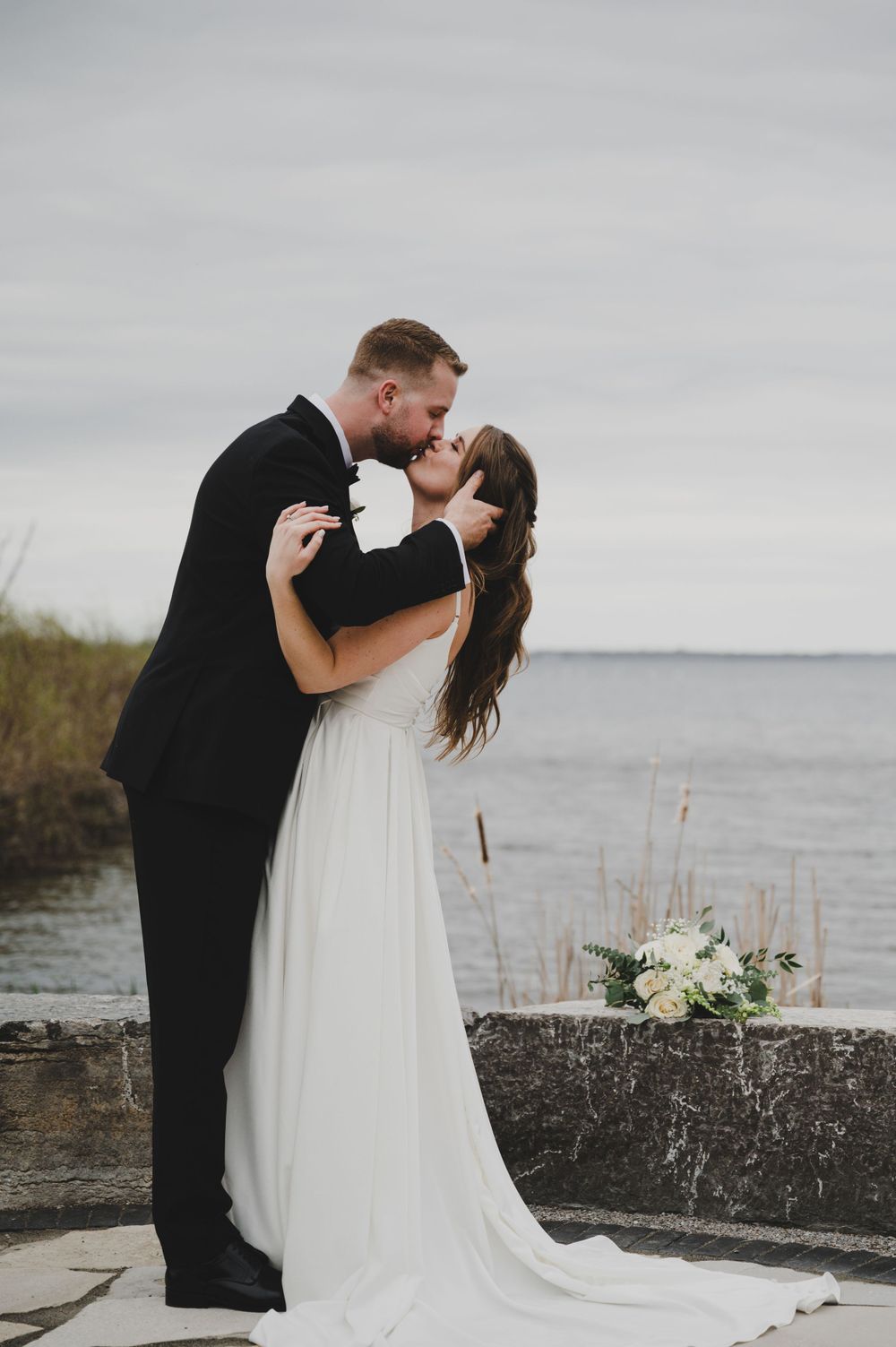 Romantic waterfront kiss with white floral bouquet in foreground.