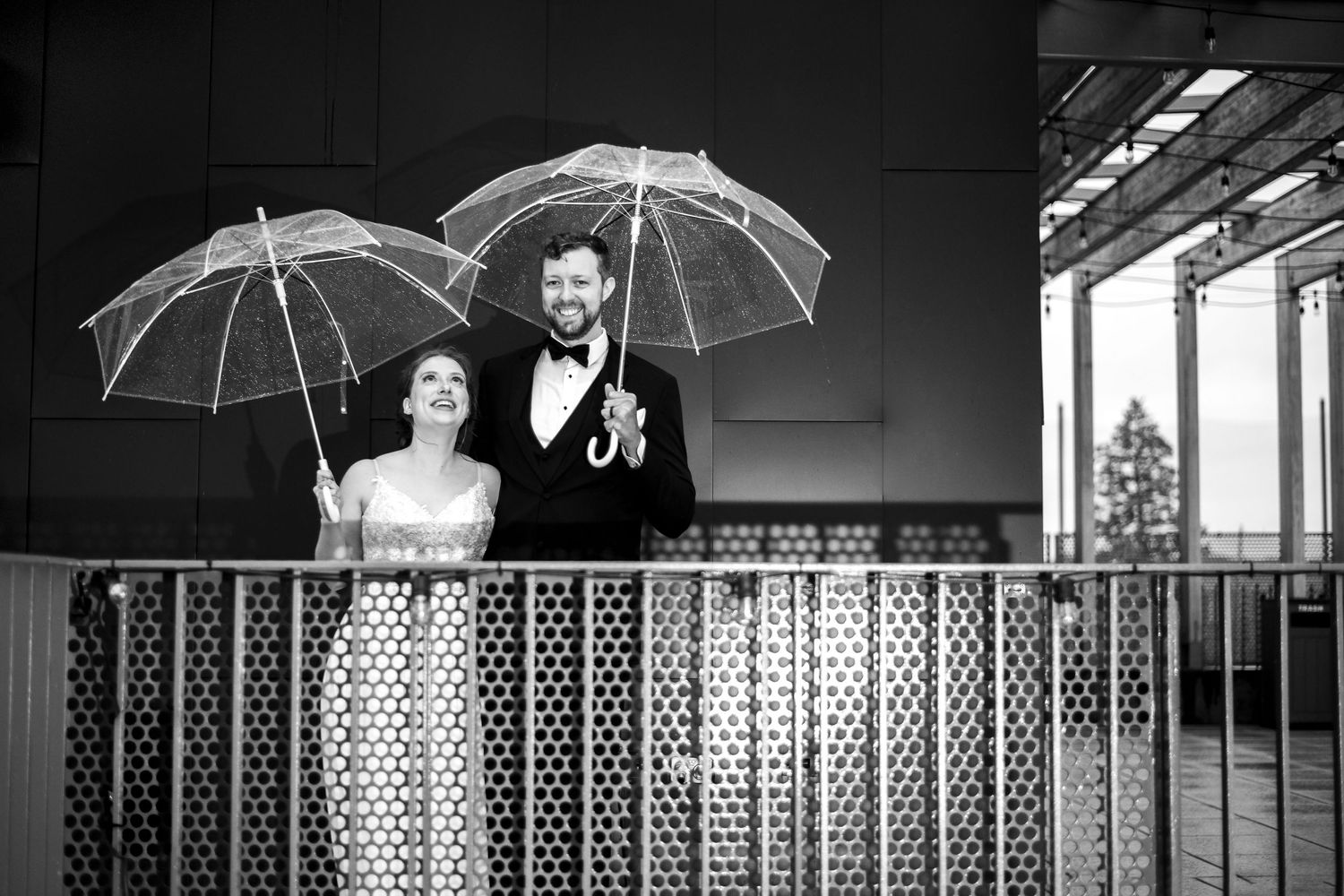 Bride and groom holding umbrellas on balcony. Rainy reception. Black and white. Romantic, editorial, authentic, classic wedding photo.