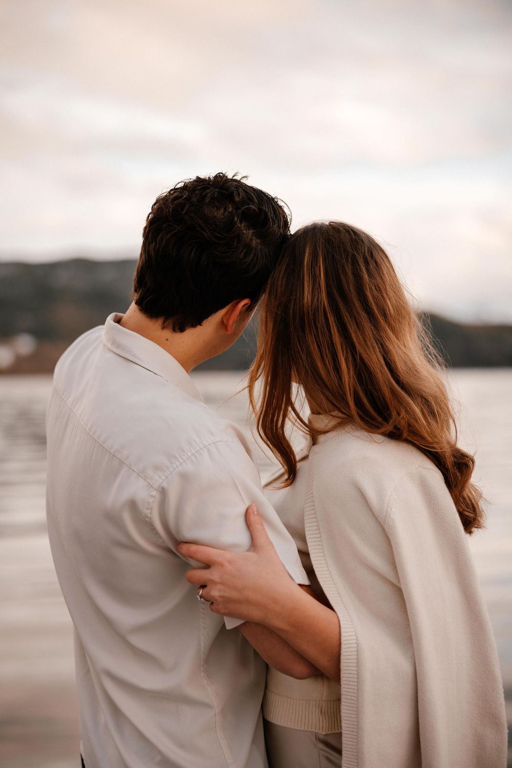 Couple in matching white outfits embrace while looking out at a waterfront view during sunset.