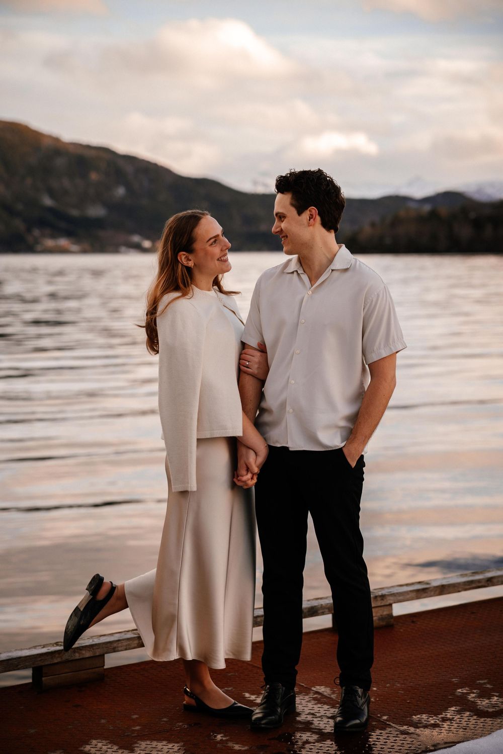 A couple shares a romantic moment on a wooden deck by a lake with mountains in the background at sunset.