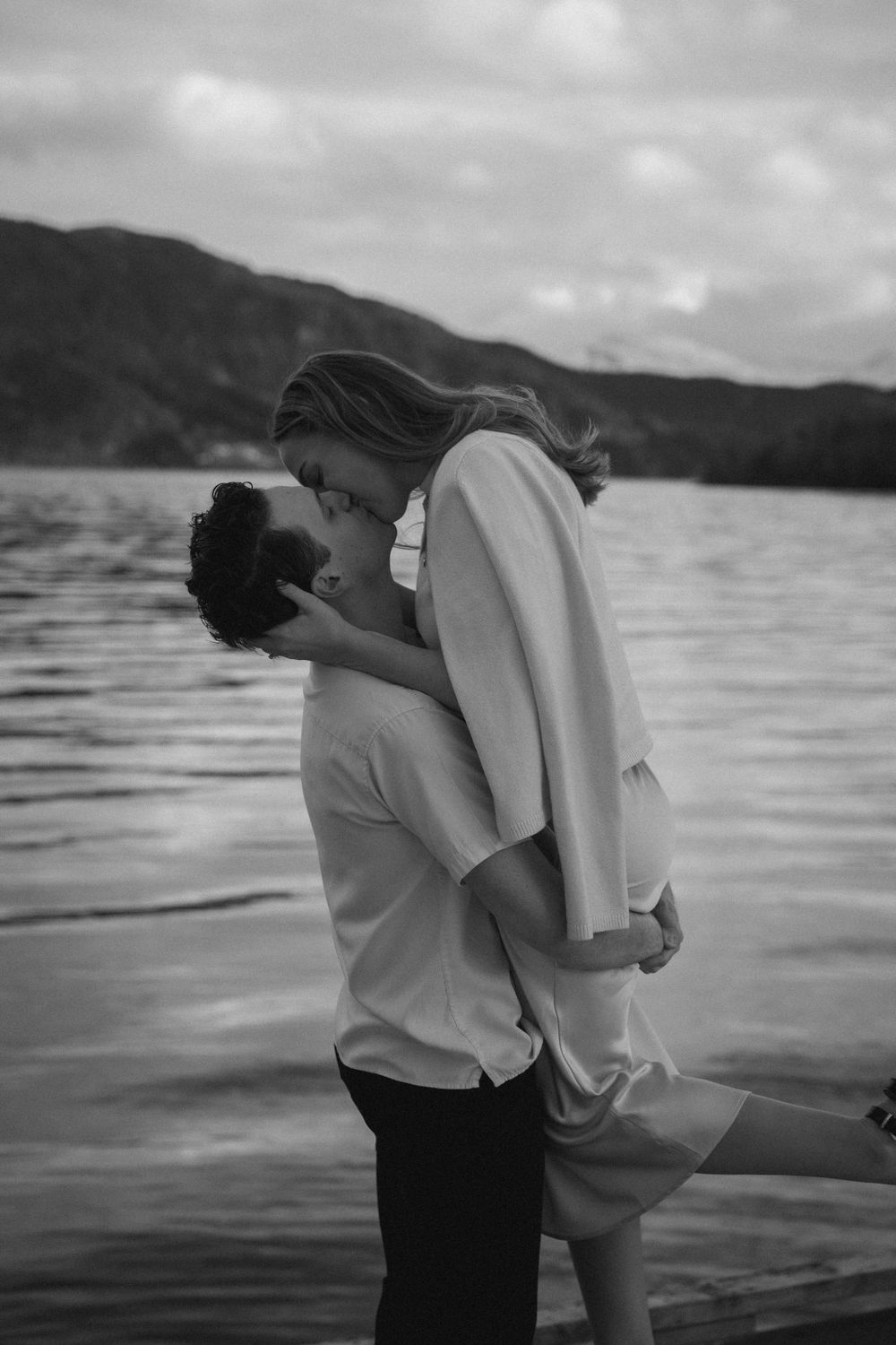 A romantic black and white moment captured by a lake with mountains in the background as a couple shares a tender kiss.