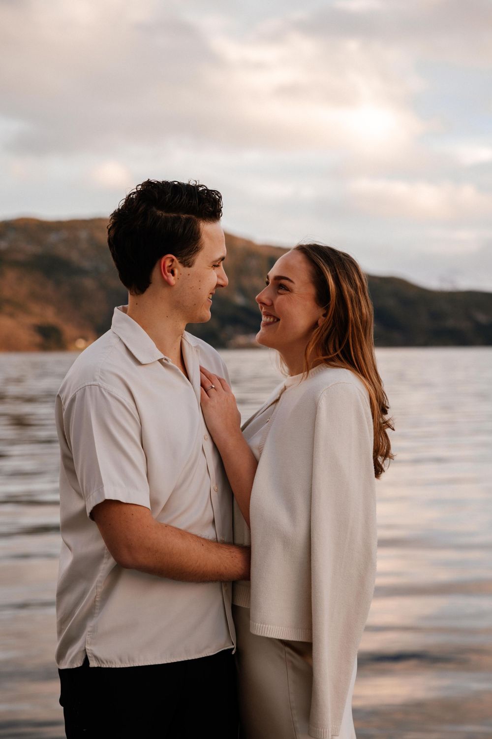 A romantic couple embraces while laughing together at sunset by a scenic lakeside with mountains in the background.
