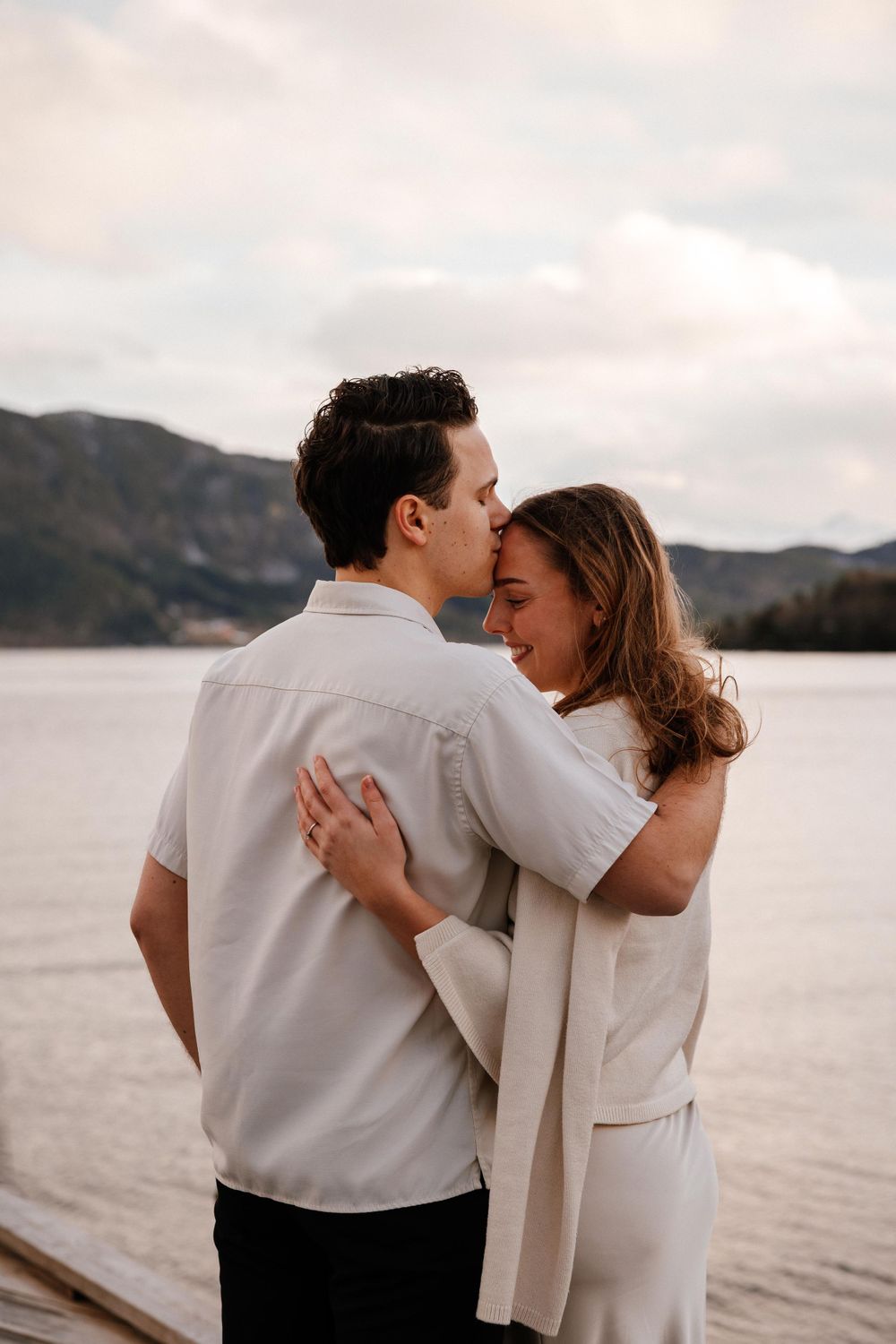 A tender romantic moment between a couple embracing by a serene lake with mountains in the background.
