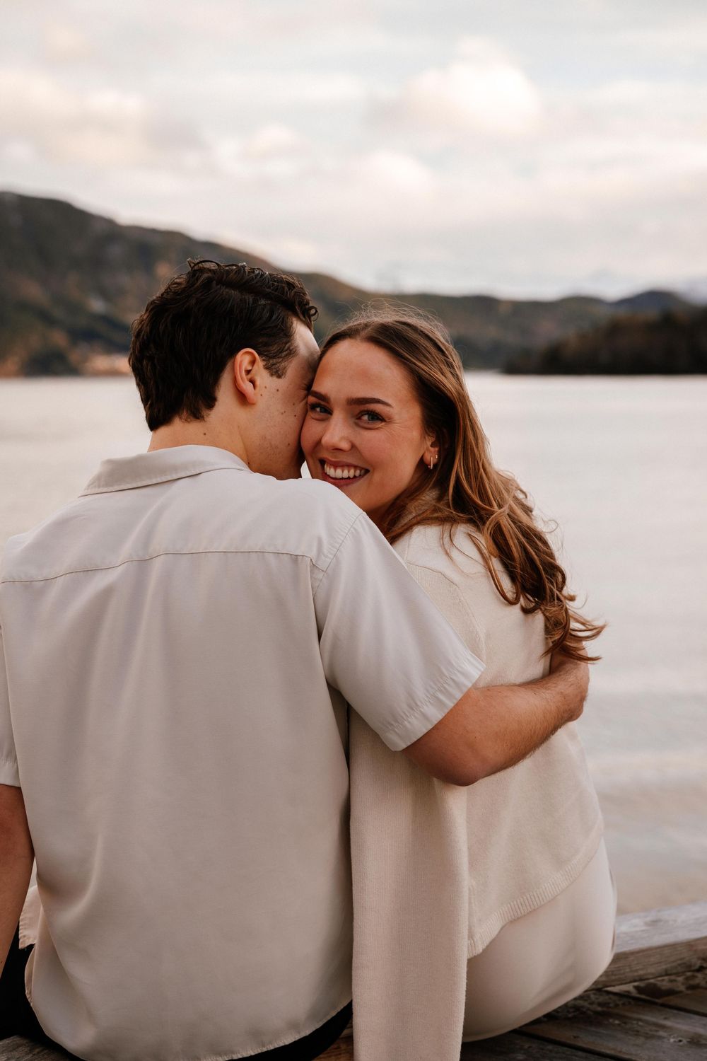 A couple in white outfits sits on a dock overlooking a serene lake with mountains in the background at sunset.