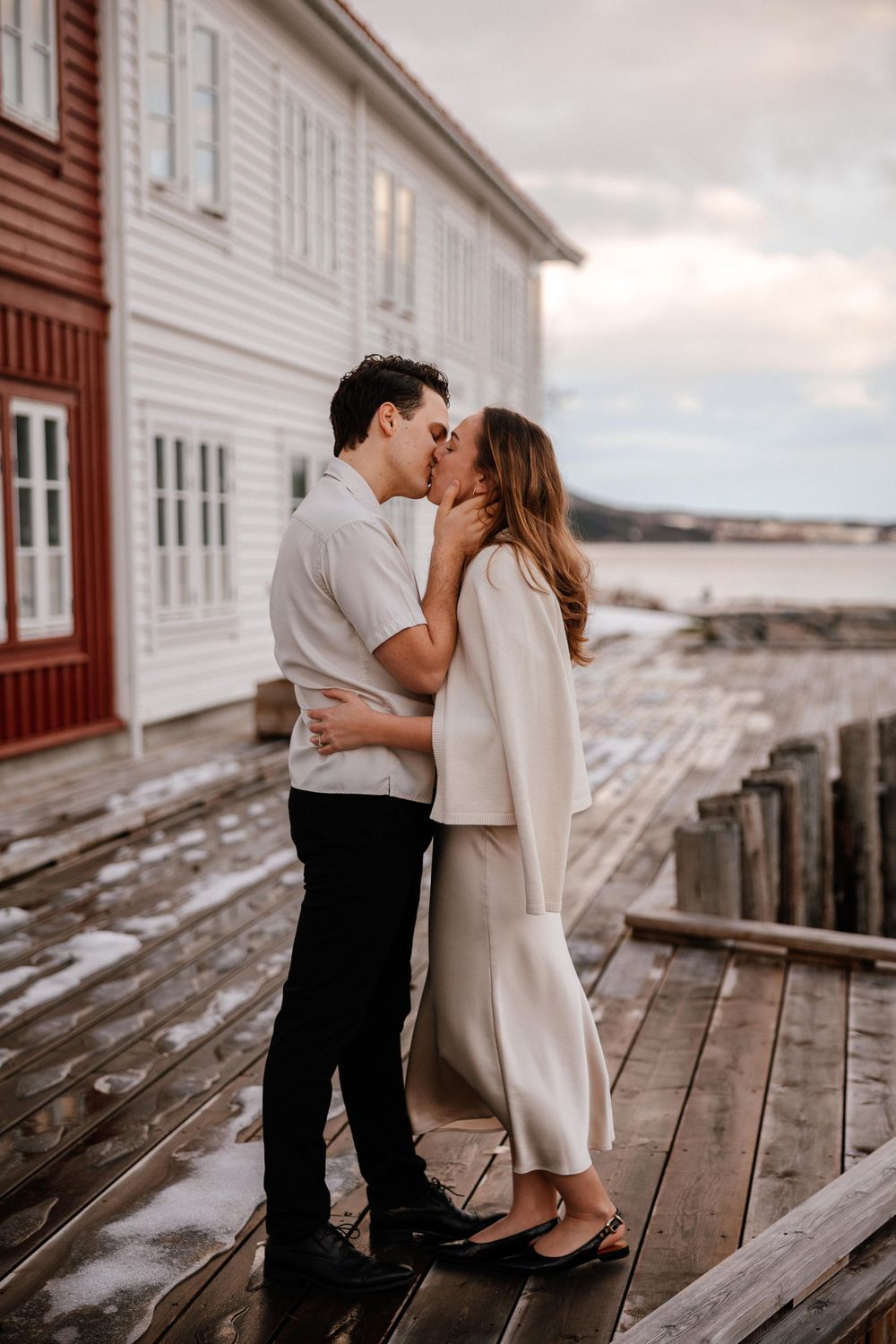 A romantic moment shared between a couple on a wooden deck by the water at sunset with rustic buildings in the background.