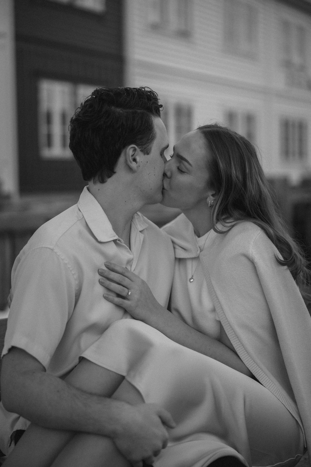 Black and white romantic photo of a couple sharing an intimate kiss while dressed in light colored clothing outdoors.