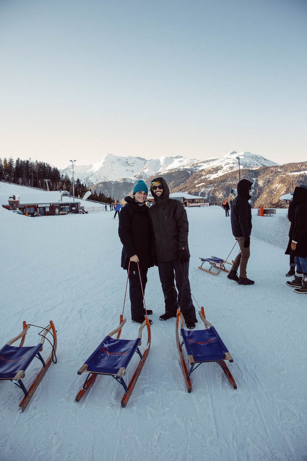 Couple posing with wooden sleds on a snowy mountain slope at a ski resort.