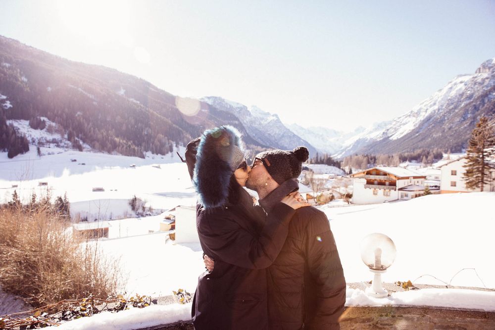 Person kissing a dog while holding it up against a stunning snowy alpine valley backdrop on a sunny winter day.
