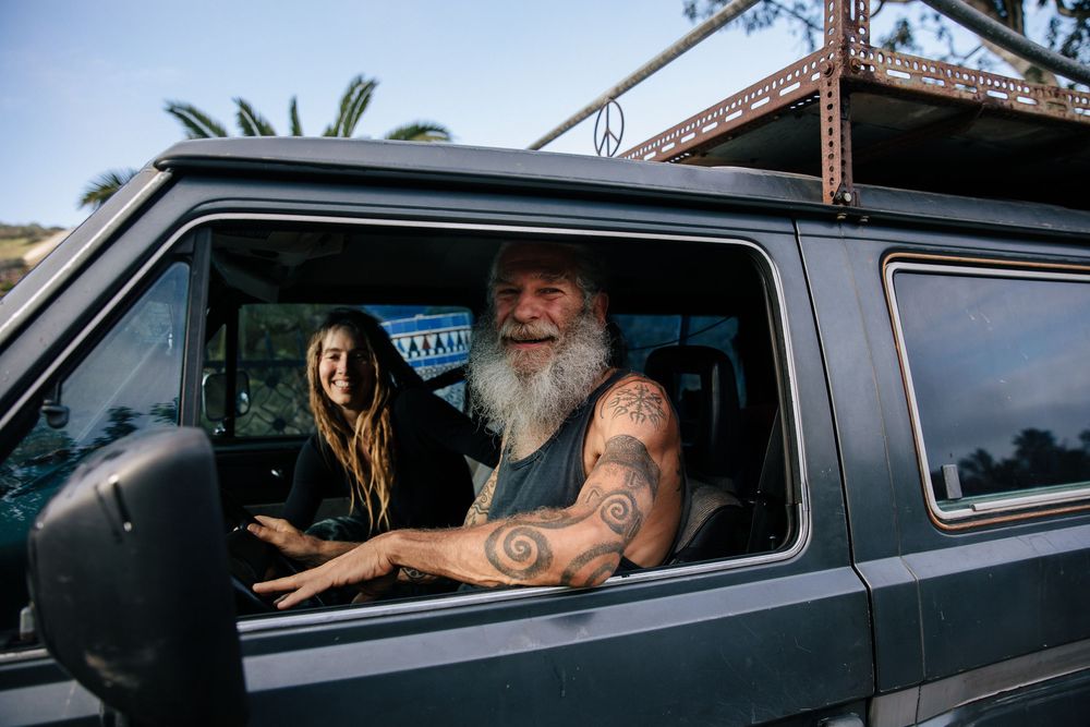 Smiling older man with white beard and spiral tattoo sitting in a van with a passenger on a sunny day.